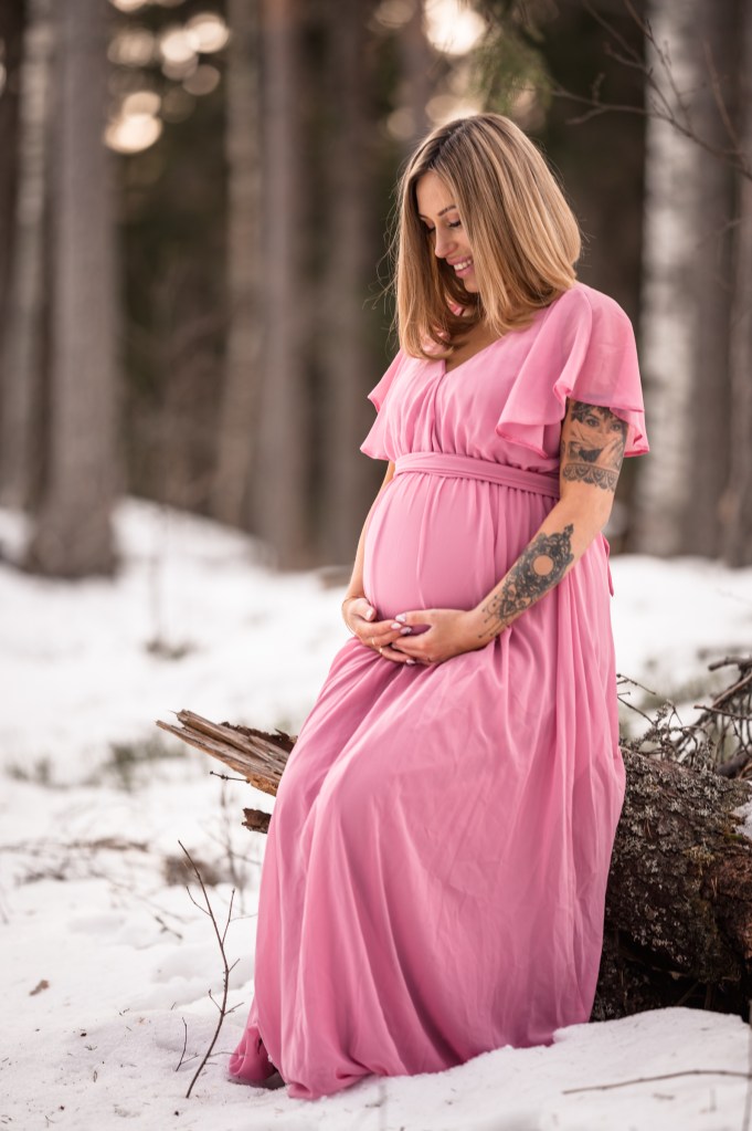 gravidporträtt taget ute i skogen där kvinnan sitter på en stubbe och håller om sin mage.
