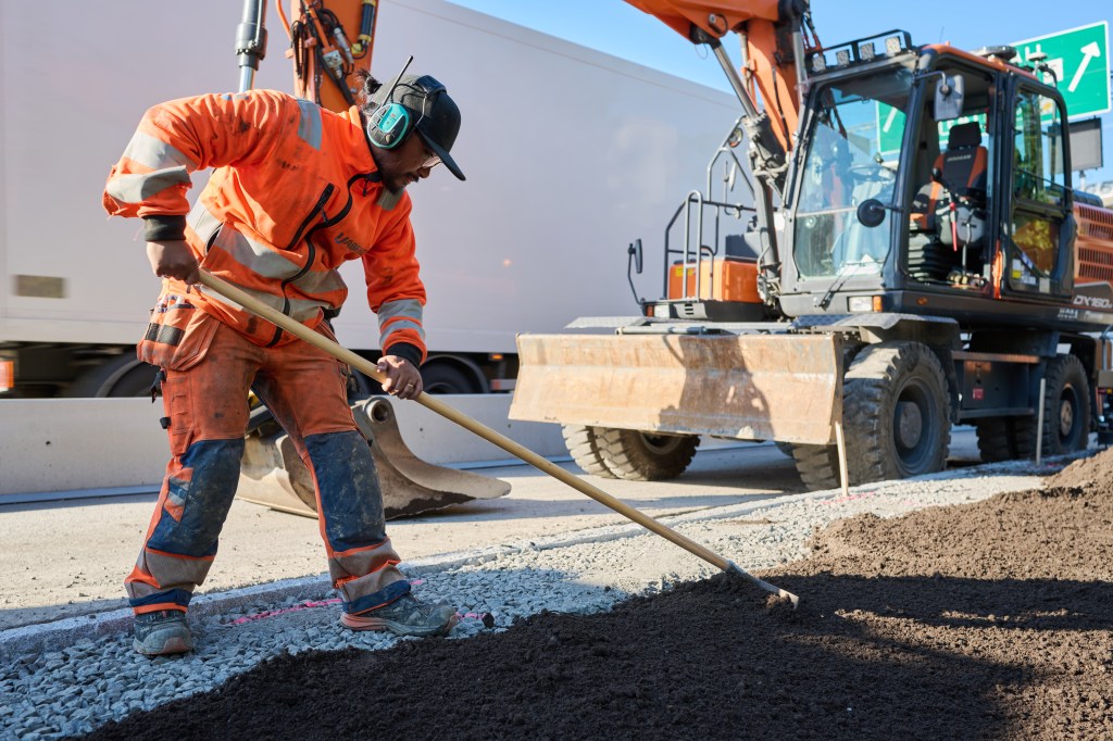 Anläggningsabetare krattar ut jord vid ett vägg bygge, grävmaskin i bakgrunden