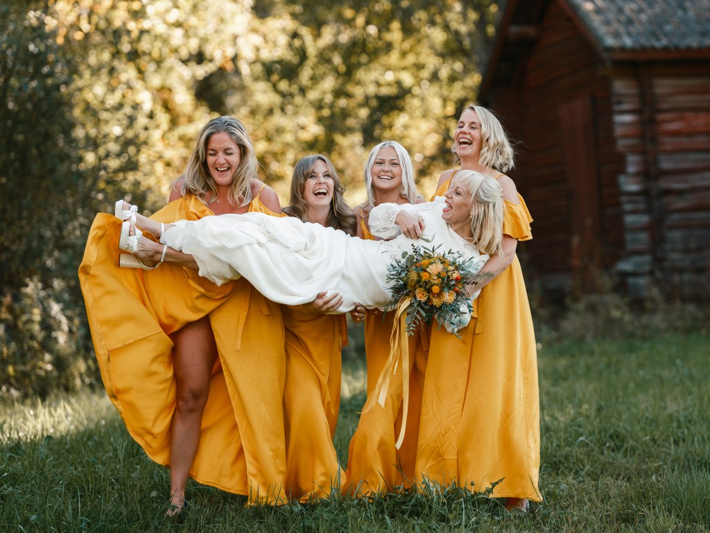 Brud som lyfts upp av sina glada tärnor i gula klänningar under en bröllopsfotografering i Järvsö, Hälsingland.