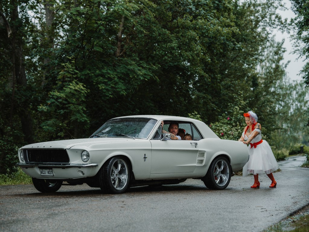 Bruden skjuter på raggarbilen medans barnen och brudgummen sitter och kör.